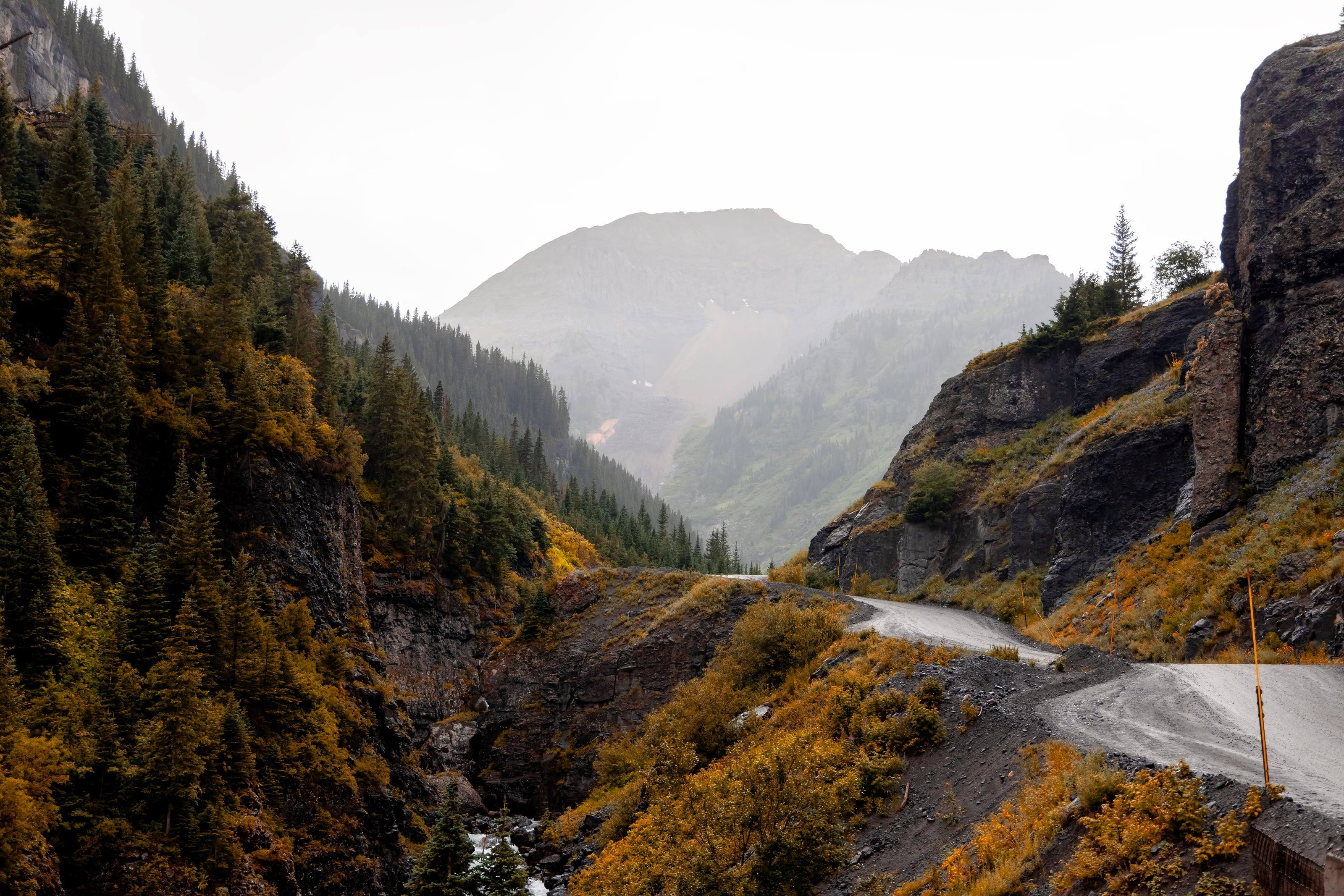A narrow shelf road cut into a cliff above a rain-washed valley in the San Juan Mountains, aspens turning below.