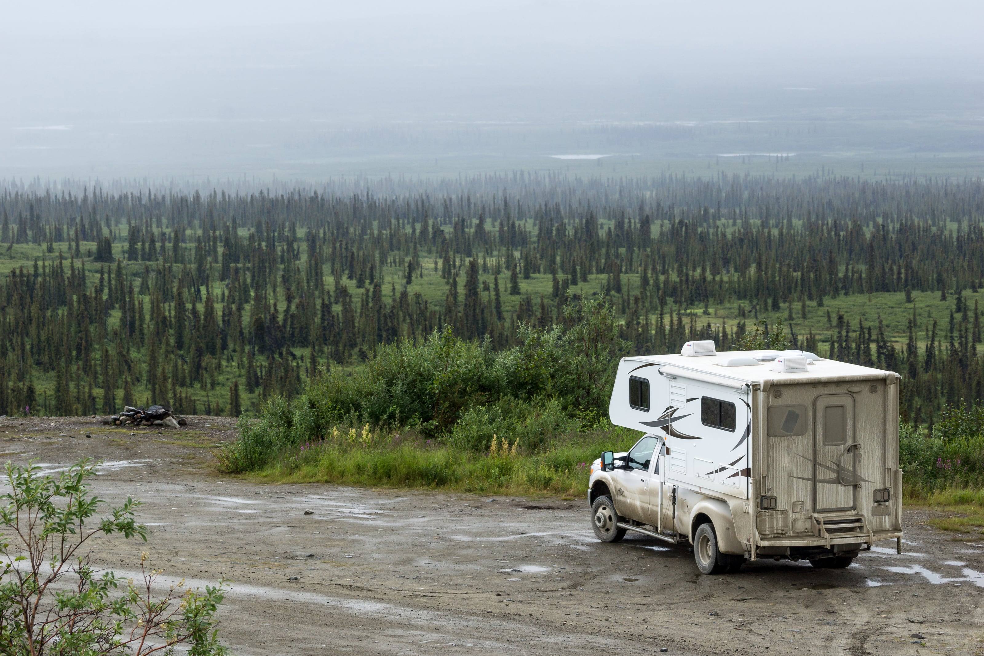 A mud-streaked pickup with a slide-in camper parked on a gravel pullout above boreal forest in the rain along the Denali Highway.