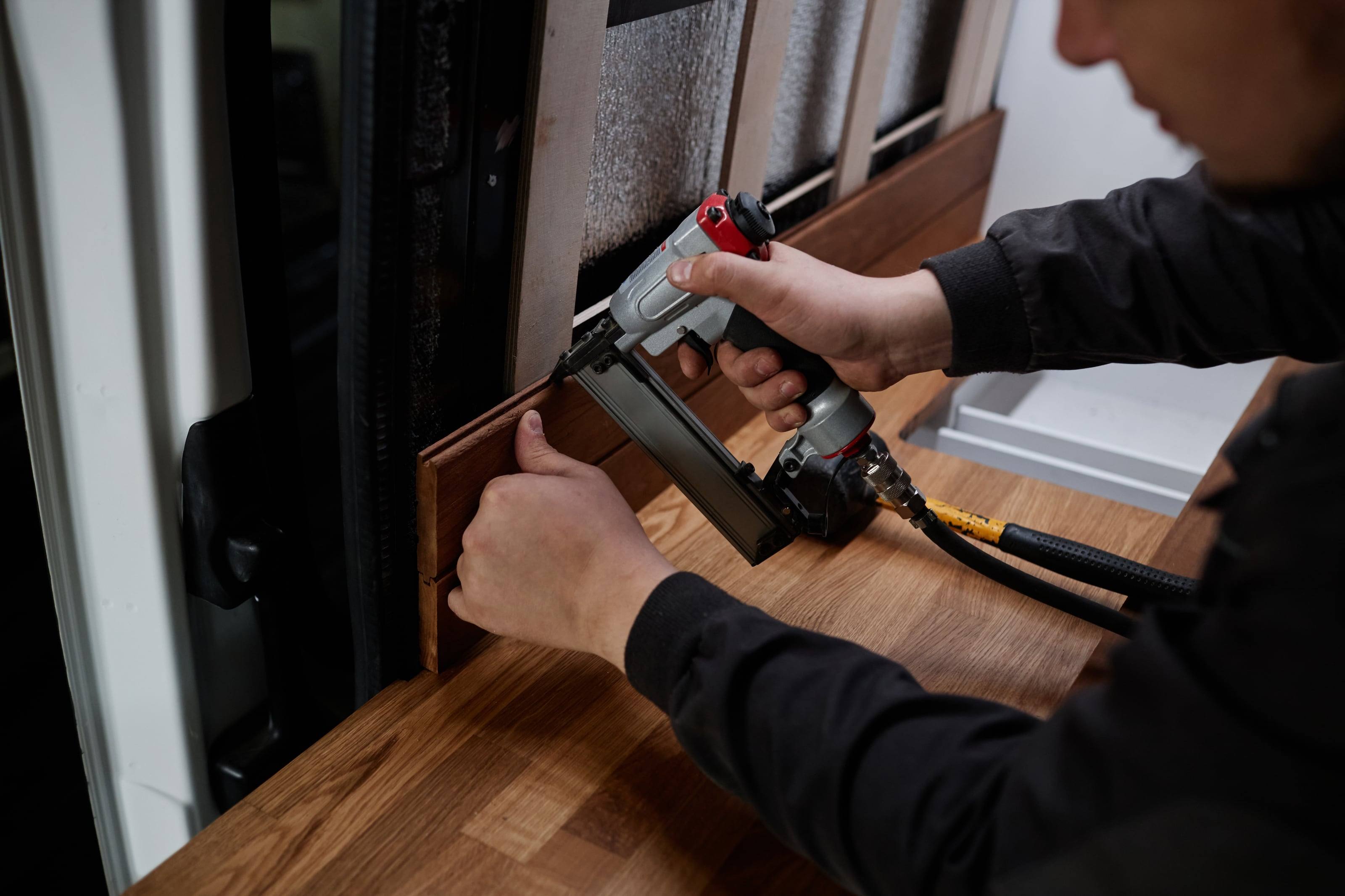 Hands fastening wooden wall boards inside a van conversion with a pneumatic nailer.