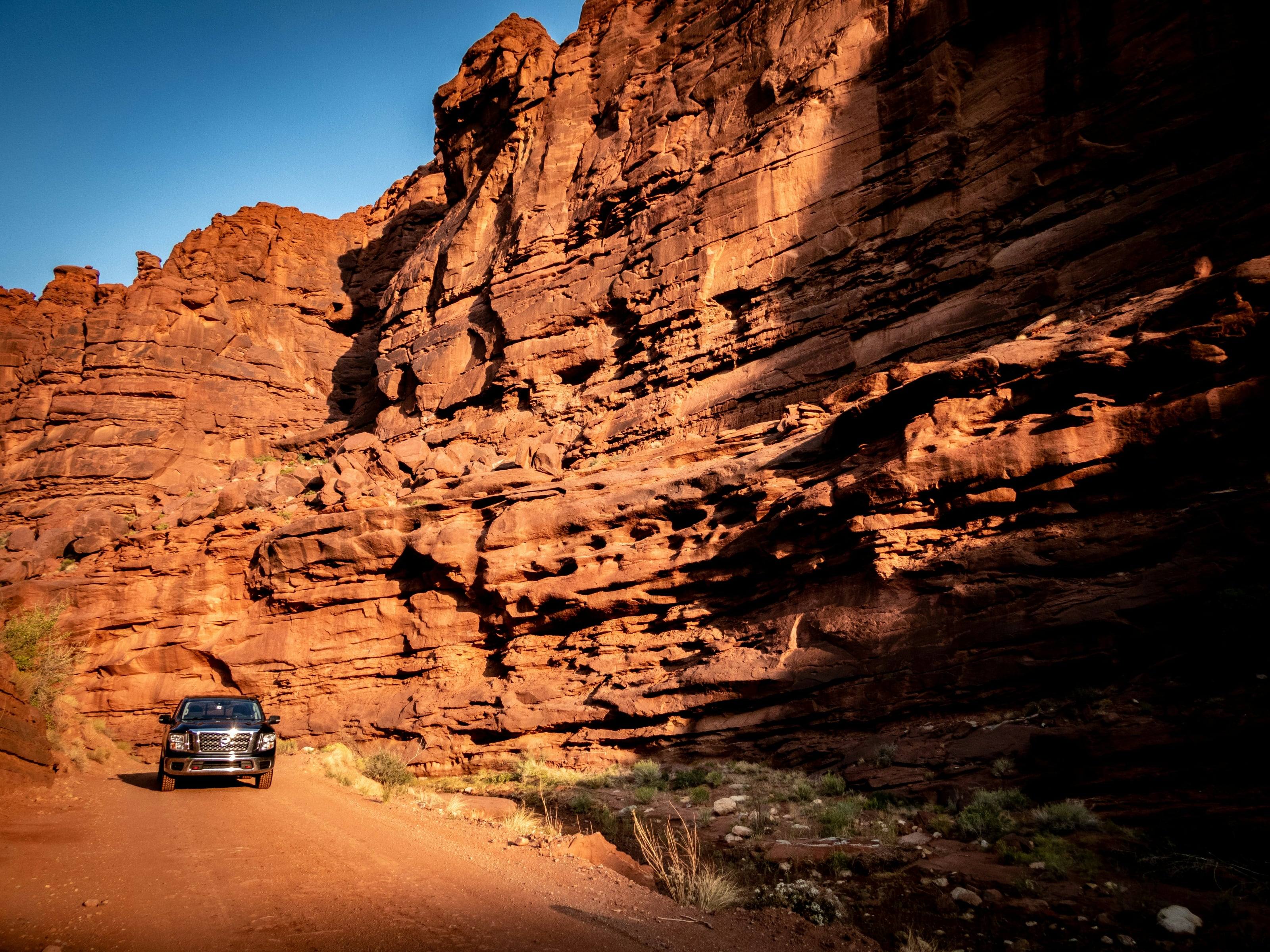 A pickup on a dirt road at the foot of a red sandstone wall lit by low sun.