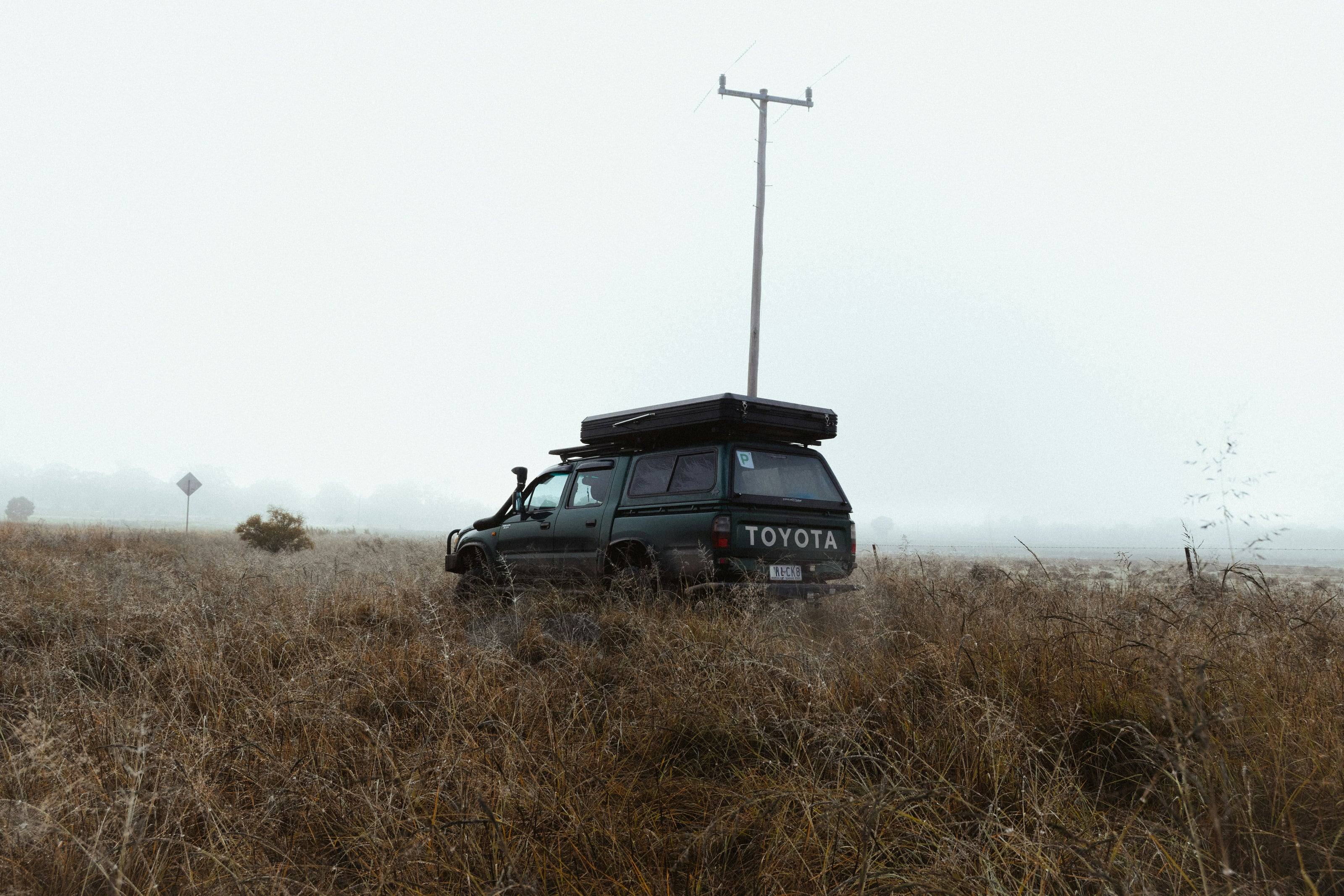 A dark green Toyota pickup with a roof tent parked in tall dry grass as mist closes in.