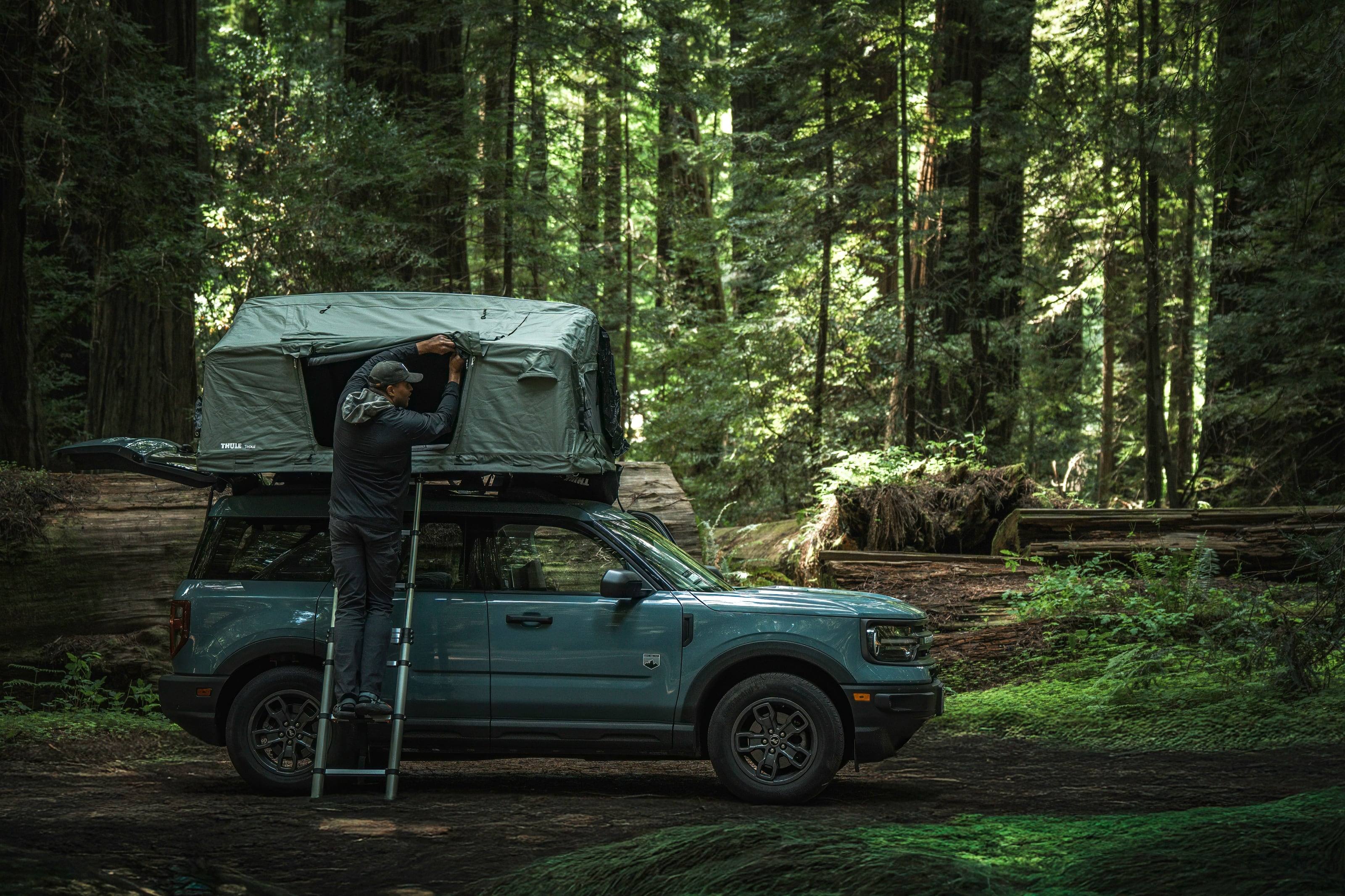 A traveler on a ladder opening the roof tent on a Bronco Sport parked in a redwood grove.