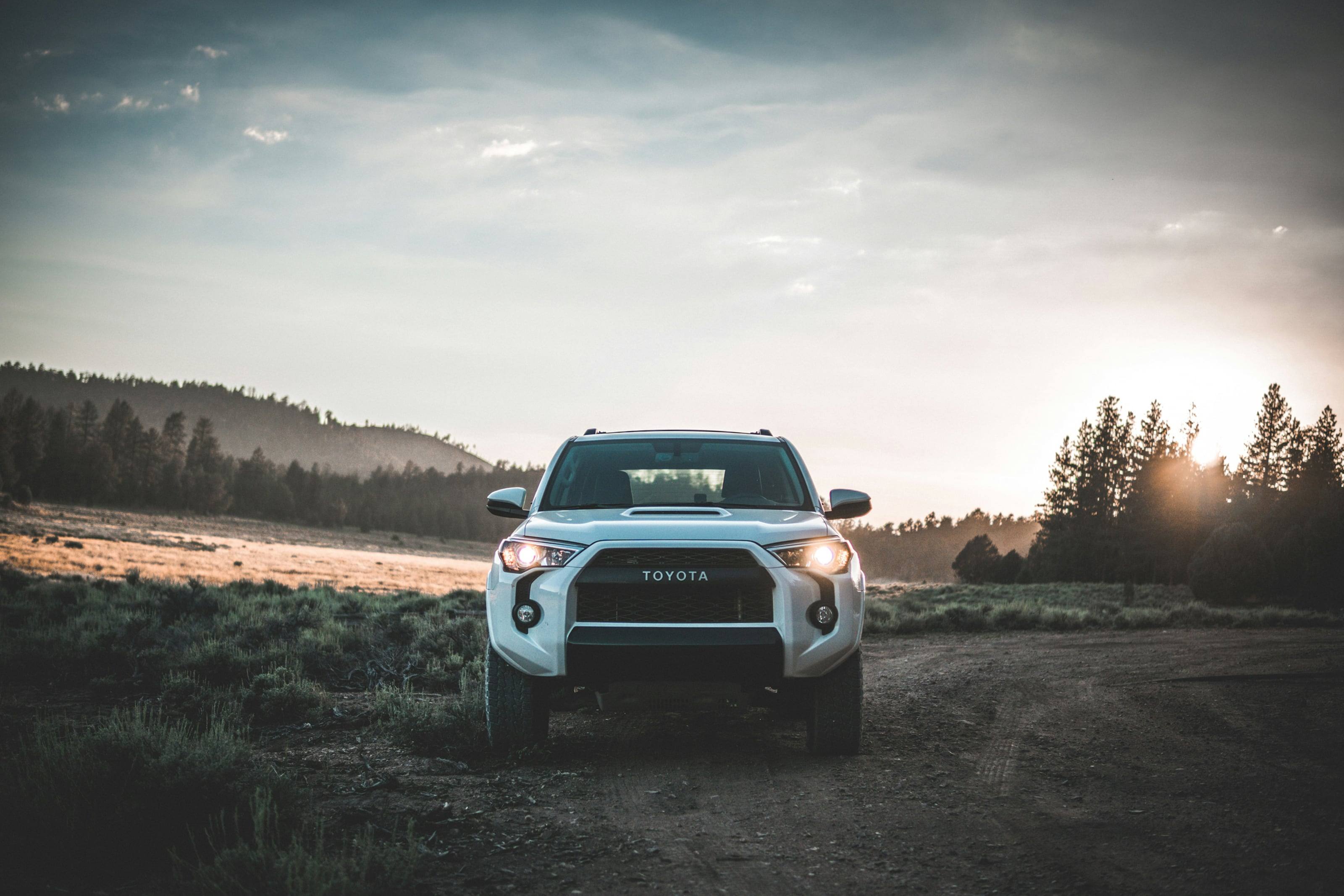 A white 4Runner on a dirt track at dusk, headlights on, pines and open ground behind.