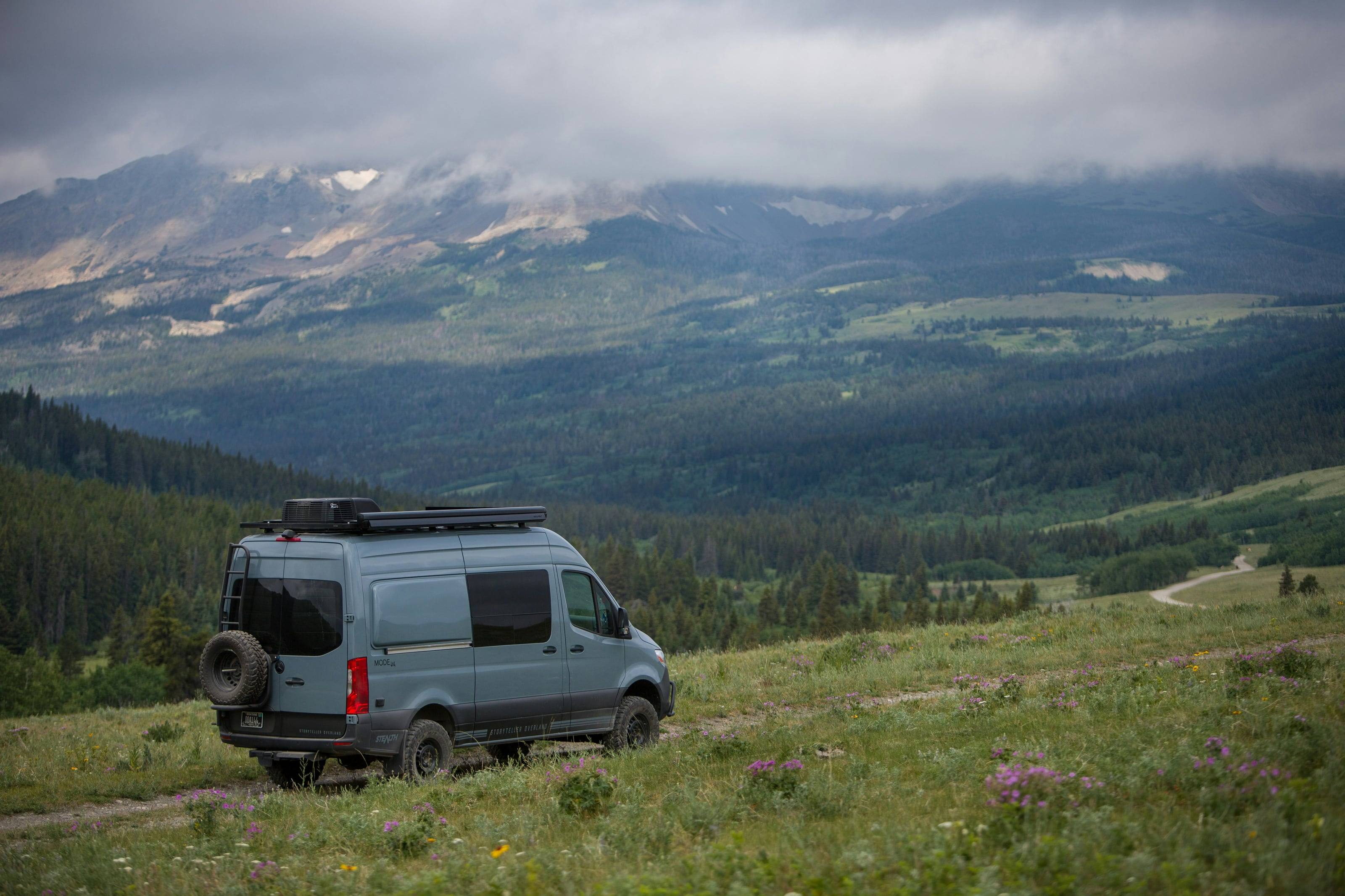 A gray 4x4 Sprinter with a roof rack on a two-track through a wildflower meadow, cloud sitting on the mountains behind.