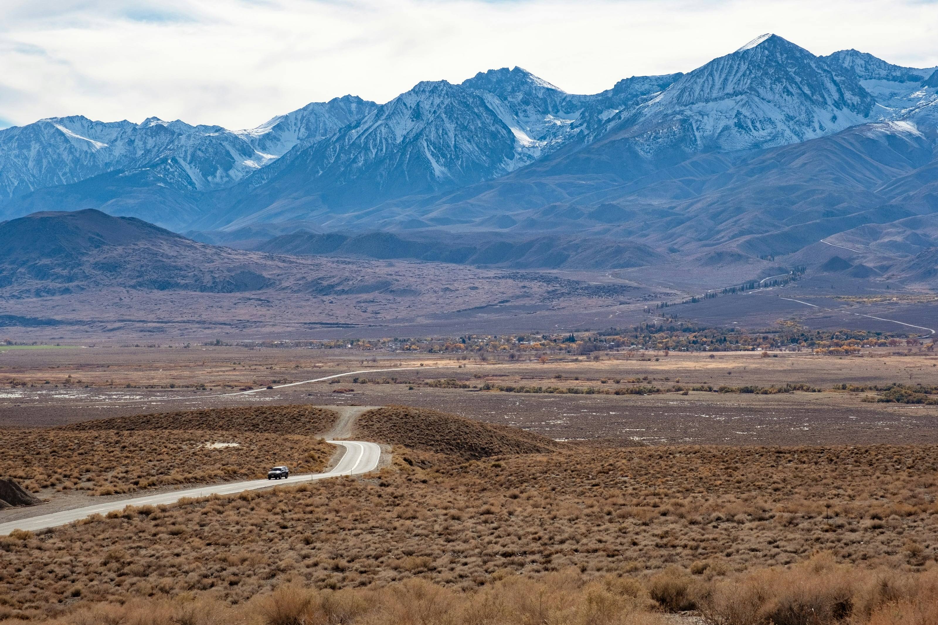 A dirt road winding across sagebrush flats toward a snow-covered mountain range, a single car far along it.
