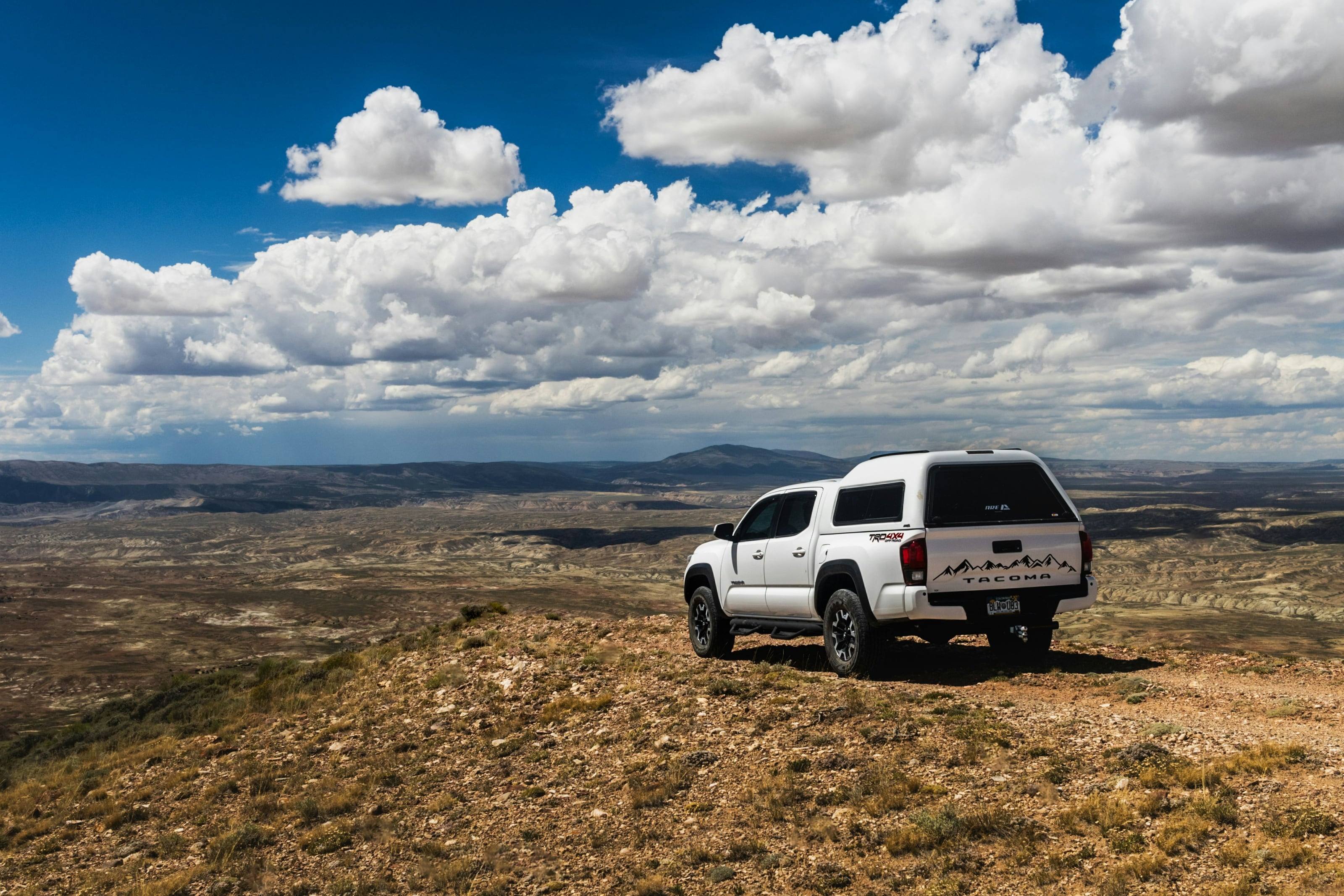 A white Tacoma with a camper shell parked on a rise above a broad, empty basin under building cloud.