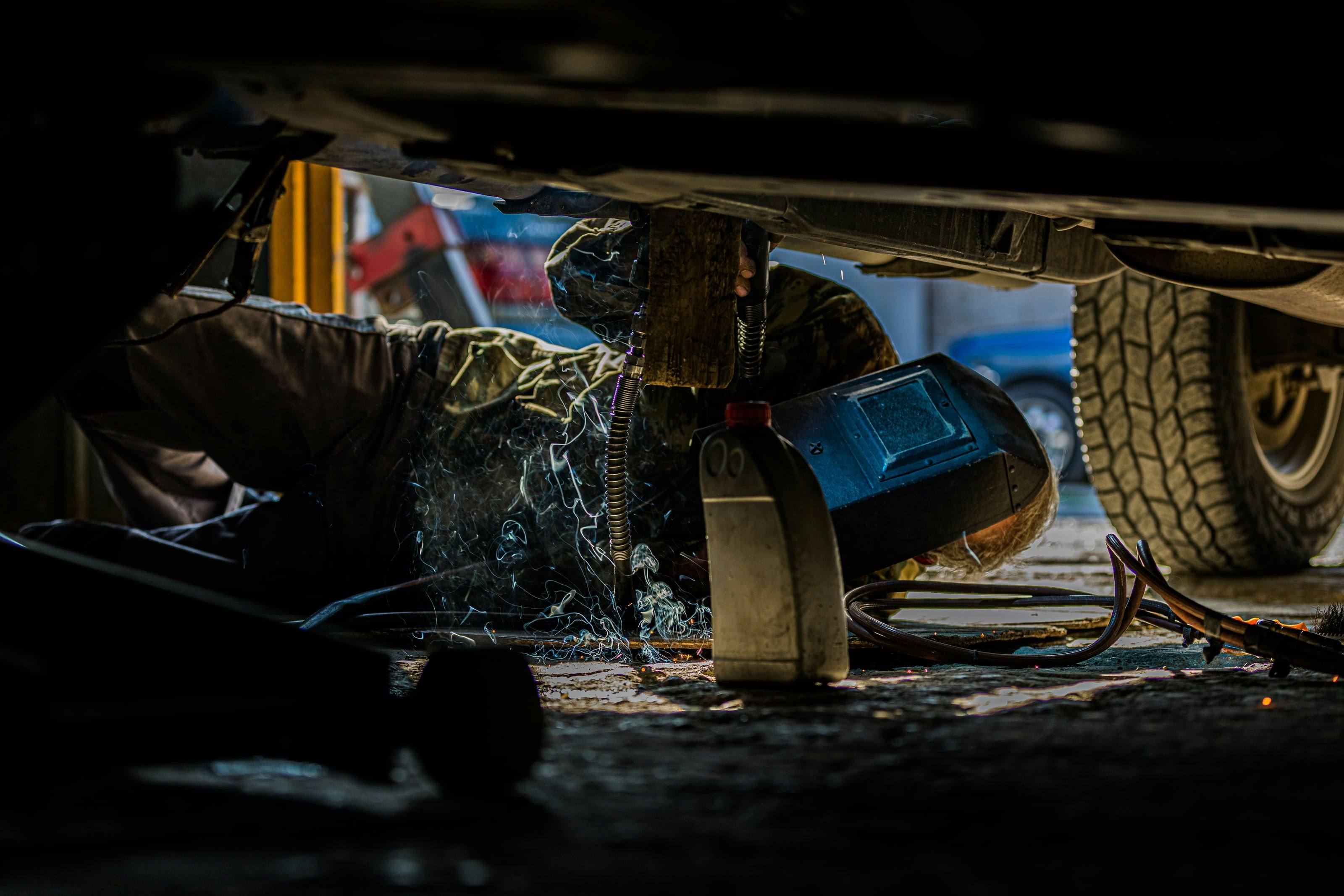 A mechanic on his back under a lifted truck, sparks flying beside an all-terrain tire.