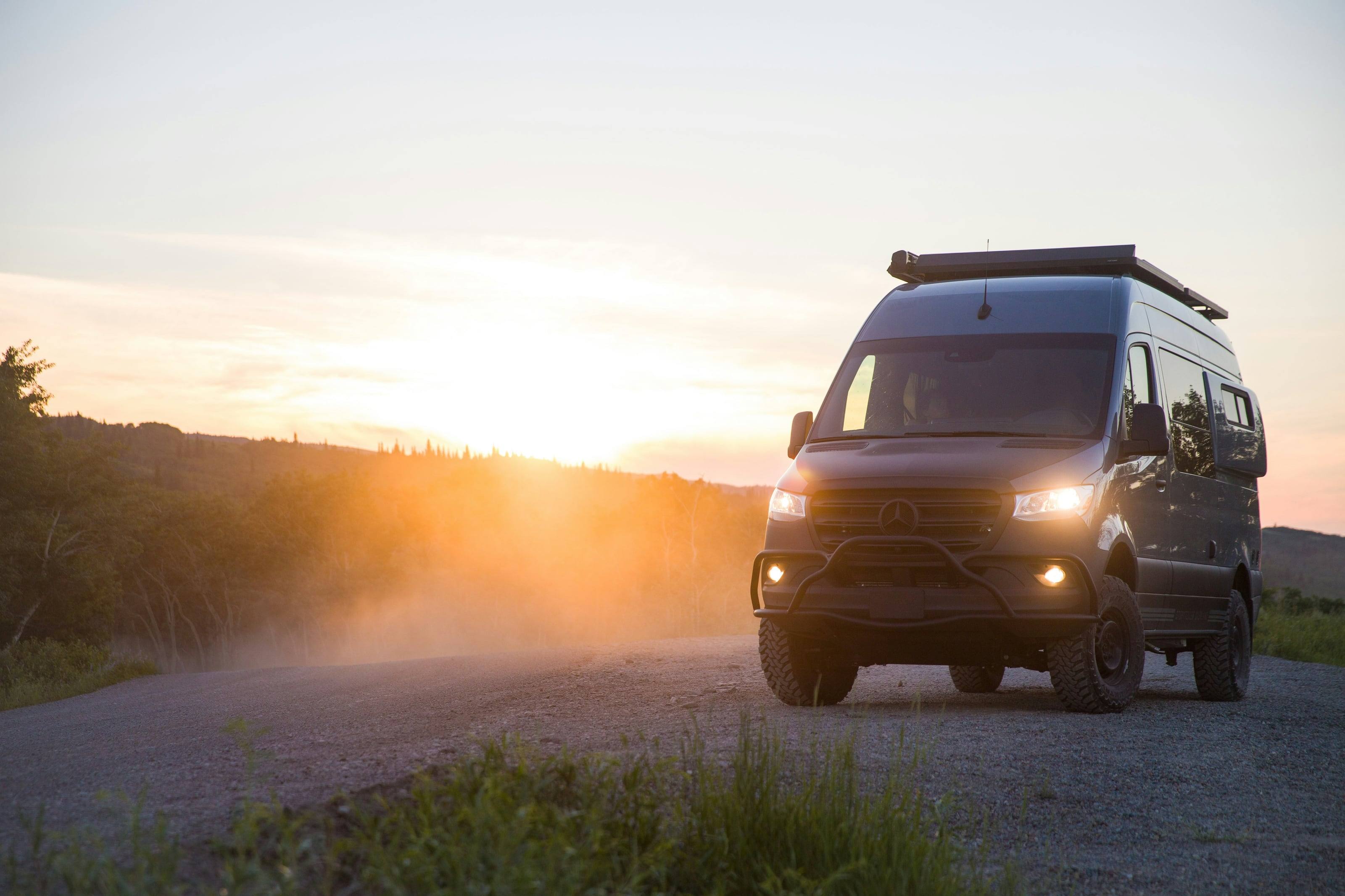 A 4x4 Sprinter van with a roof rack on a gravel road, dust lit by the low sun behind it.