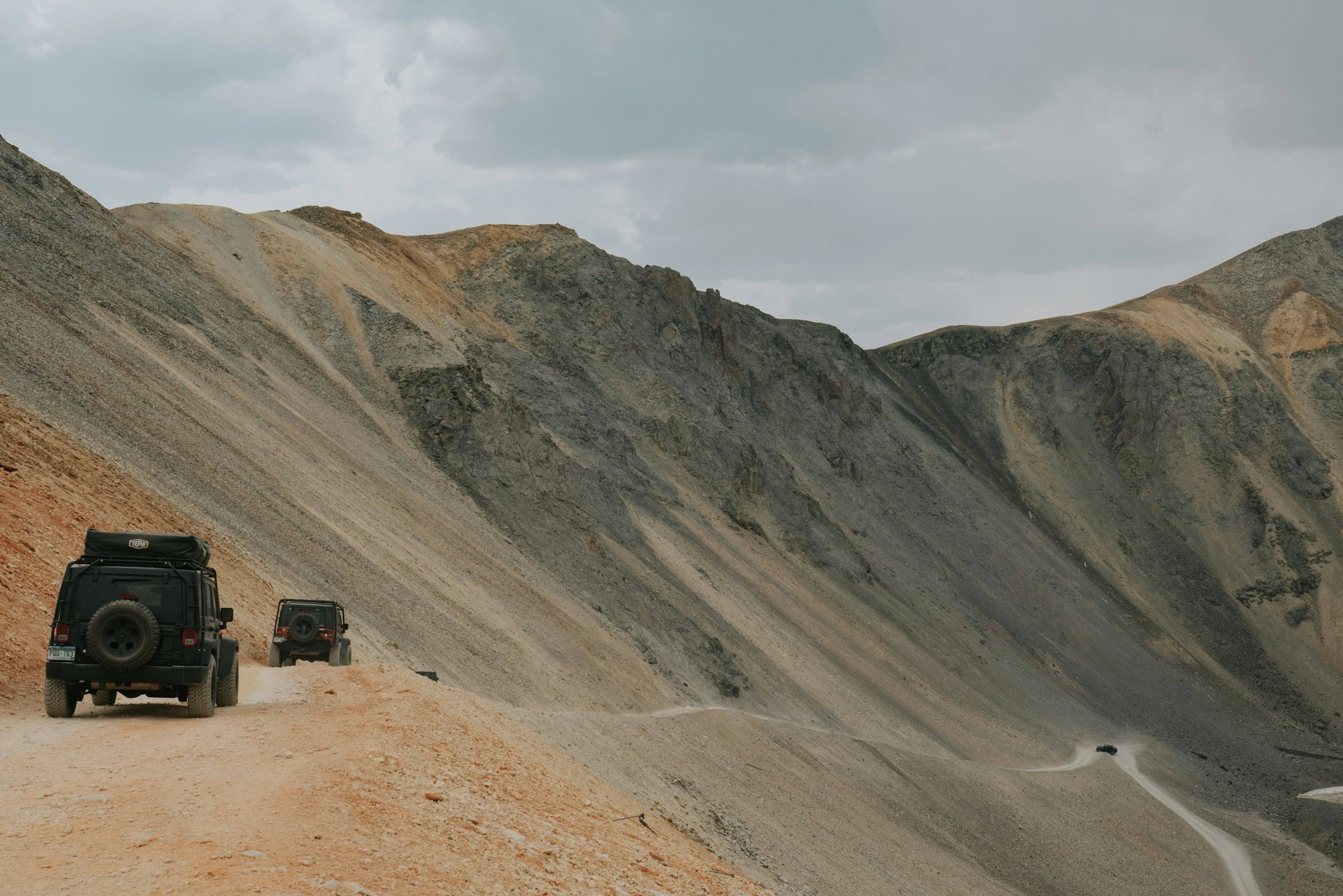 Two 4x4s with roof tents climbing a switchback shelf road across a high scree slope under low cloud.