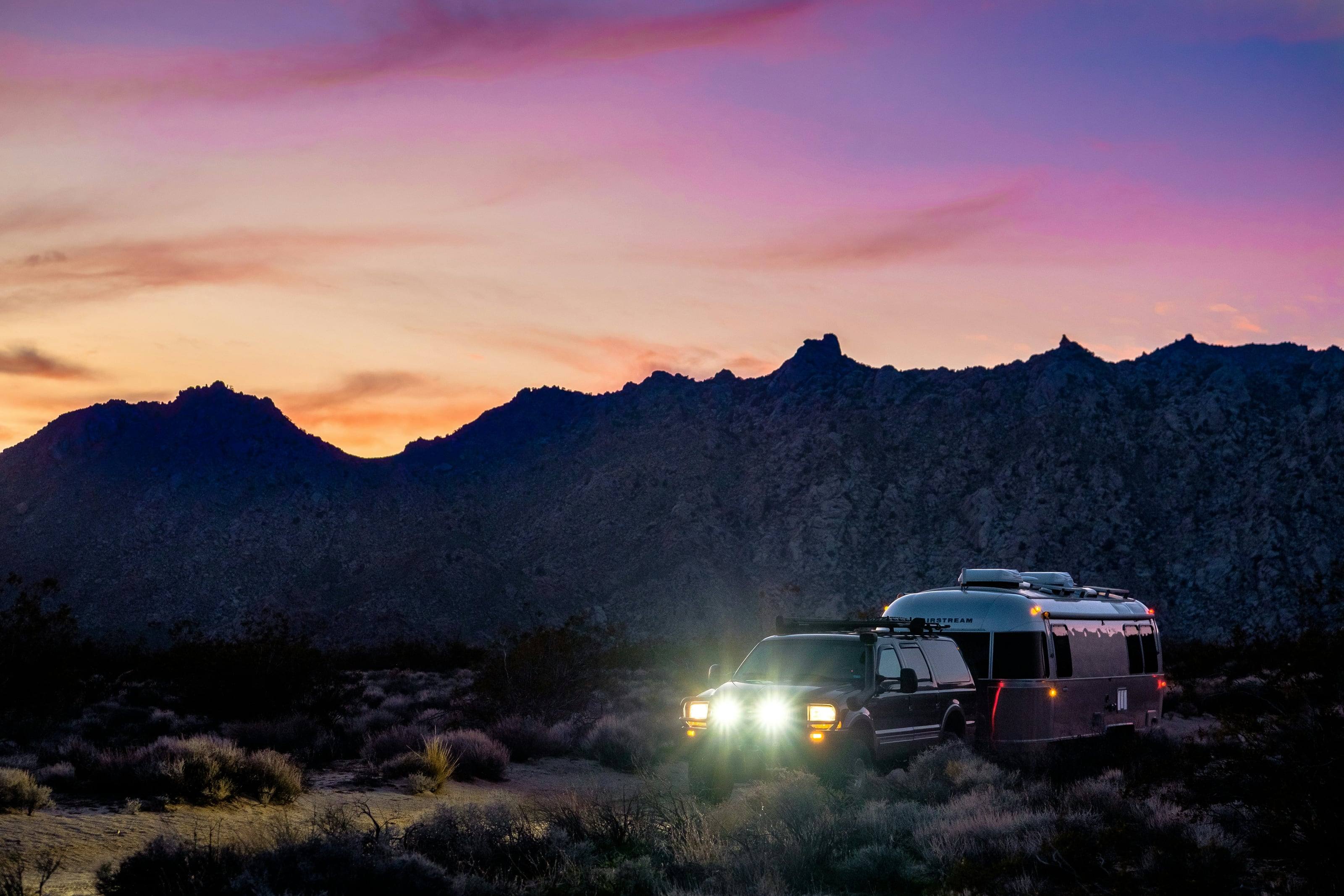 A 4Runner towing an off-road trailer along a desert track at last light, headlights on below a jagged ridge.