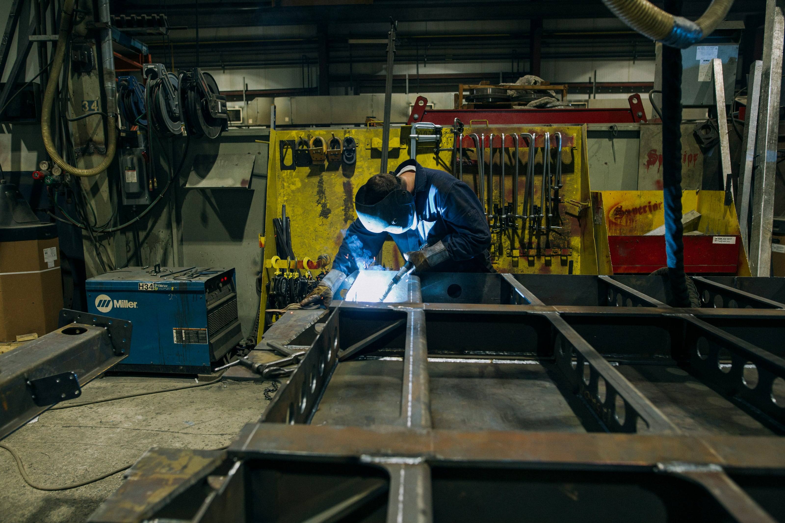 A welder joining the rails of a bare steel chassis in a fabrication shop, tool board and welder behind.